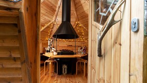 Interior acogedor de una cabaña de madera con una mesa central, una chimenea negra y luces colgantes.