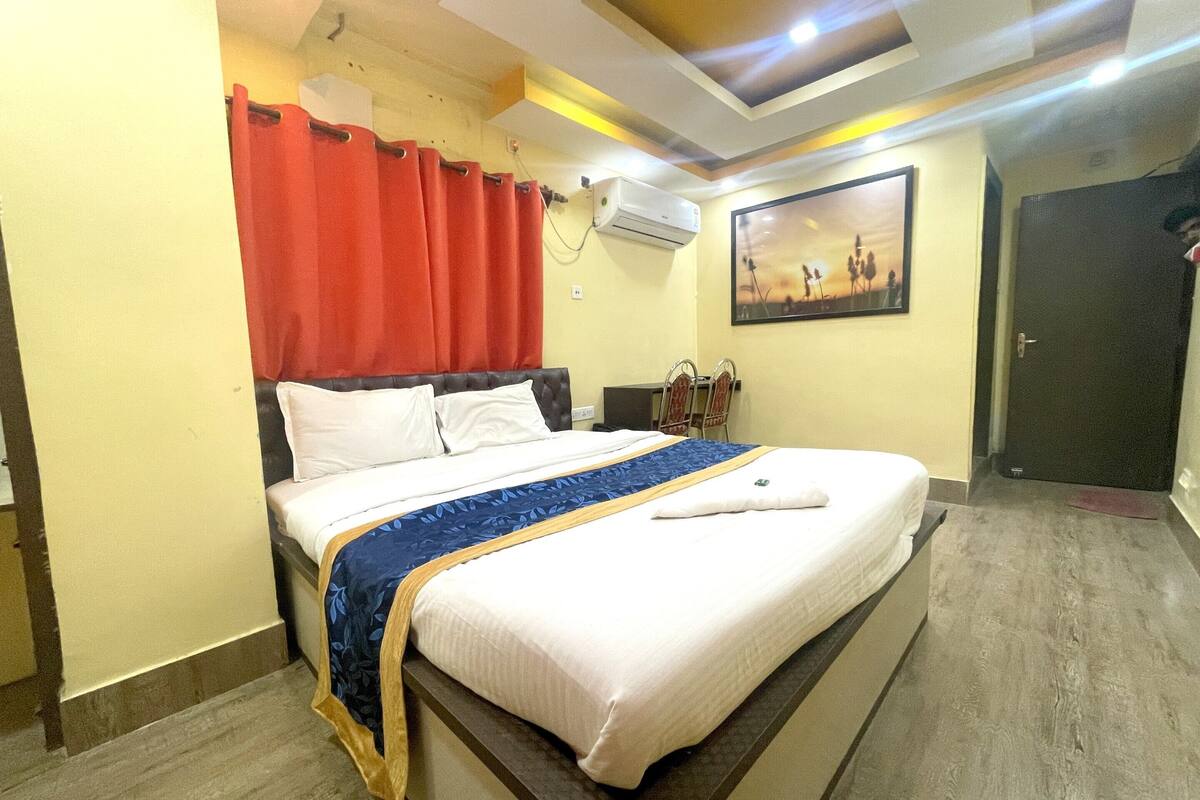 A hotel room with a bed, a red curtain, an air conditioning unit, a painting on the wall, and a door.