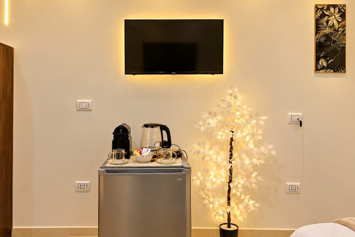 A hotel room with a flat-screen TV mounted on the wall, a minibar with a coffee maker, and a decorative artificial tree.