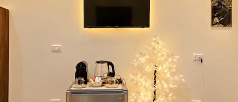 A hotel room with a flat-screen TV mounted on the wall, a minibar with a coffee maker, and a decorative artificial tree.