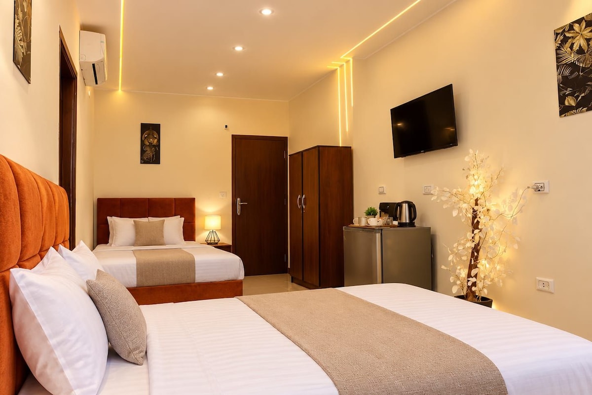 A hotel room with two beds, a television, a minibar, and a decorative plant.