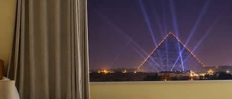A hotel room with two beds, a window with a view of a pyramid at night, and a lamp on the left.