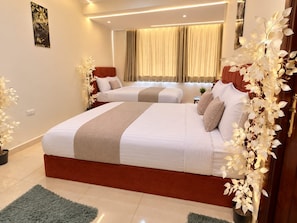 A hotel room with two beds, a large window with curtains, a decorative plant, and a wall-mounted artwork.