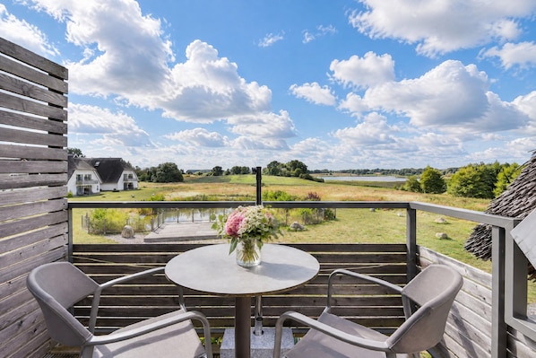 Une terrasse en bois avec une table ronde et deux chaises, d’où l’on peut admirer un paysage rural avec un bâtiment blanc et une étendue d’eau.