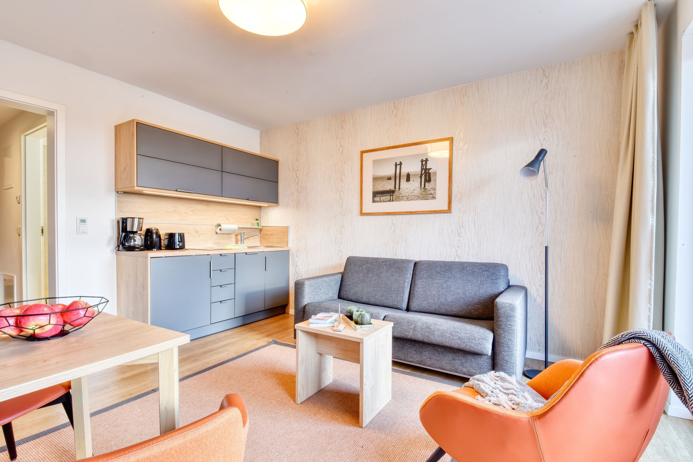 Un salon moderne avec un canapé gris, un fauteuil orange et une table basse en bois. On aperçoit à l’arrière-plan un coin cuisine avec des placards gris et un plan de travail en bois.