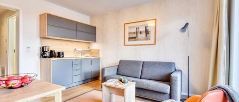 Un salon moderne avec un canapé gris, un fauteuil orange et une table basse en bois. On aperçoit à l’arrière-plan un coin cuisine avec des placards gris et un plan de travail en bois.
