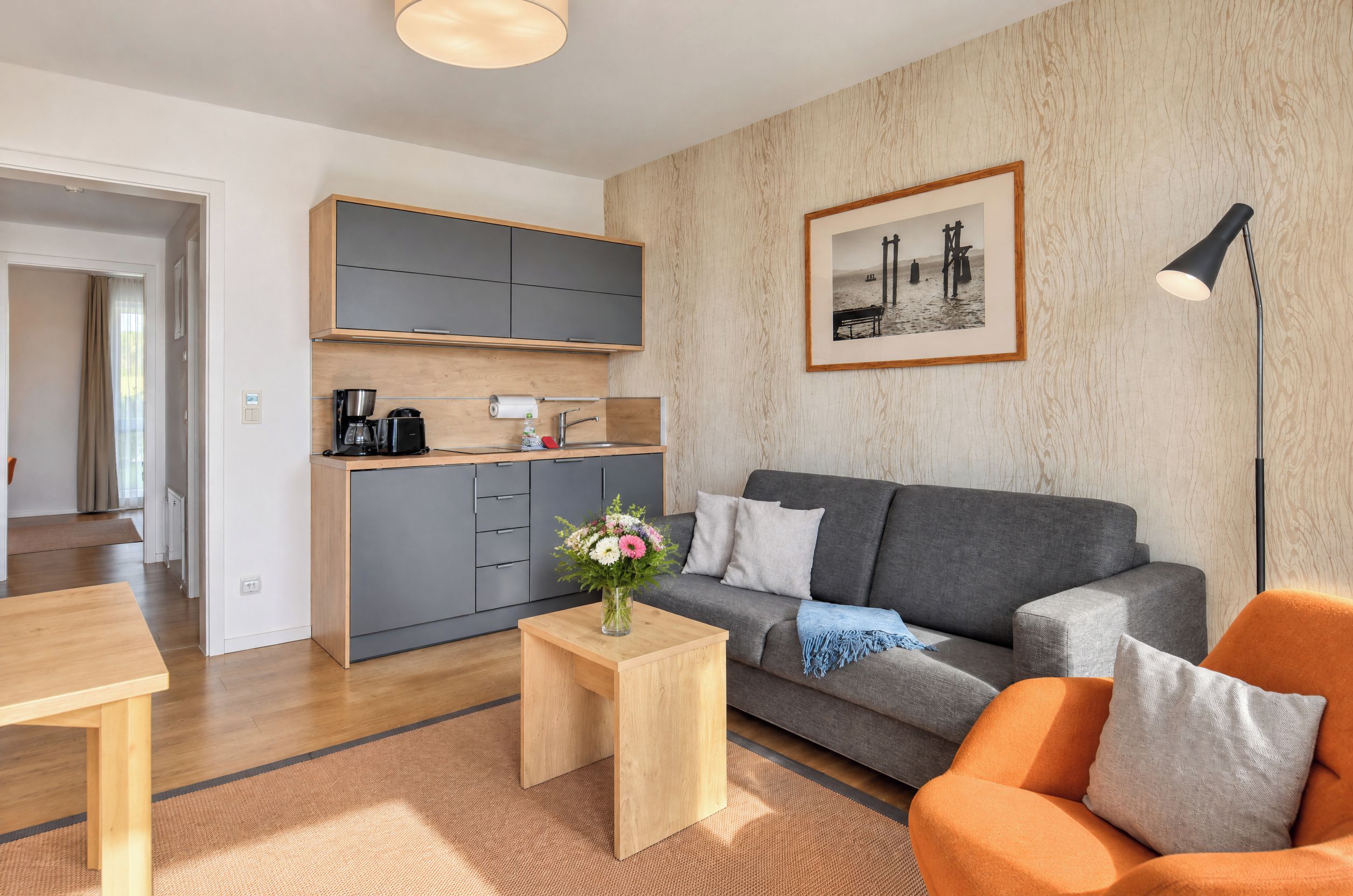 A modern living room with a grey sofa, a wooden coffee table with a vase of flowers, a small kitchen area with a coffee machine, and a framed picture on the wall.