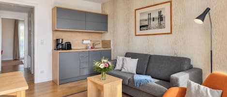 A modern living room with a grey sofa, a wooden coffee table with a vase of flowers, a small kitchen area with a coffee machine, and a framed picture on the wall.
