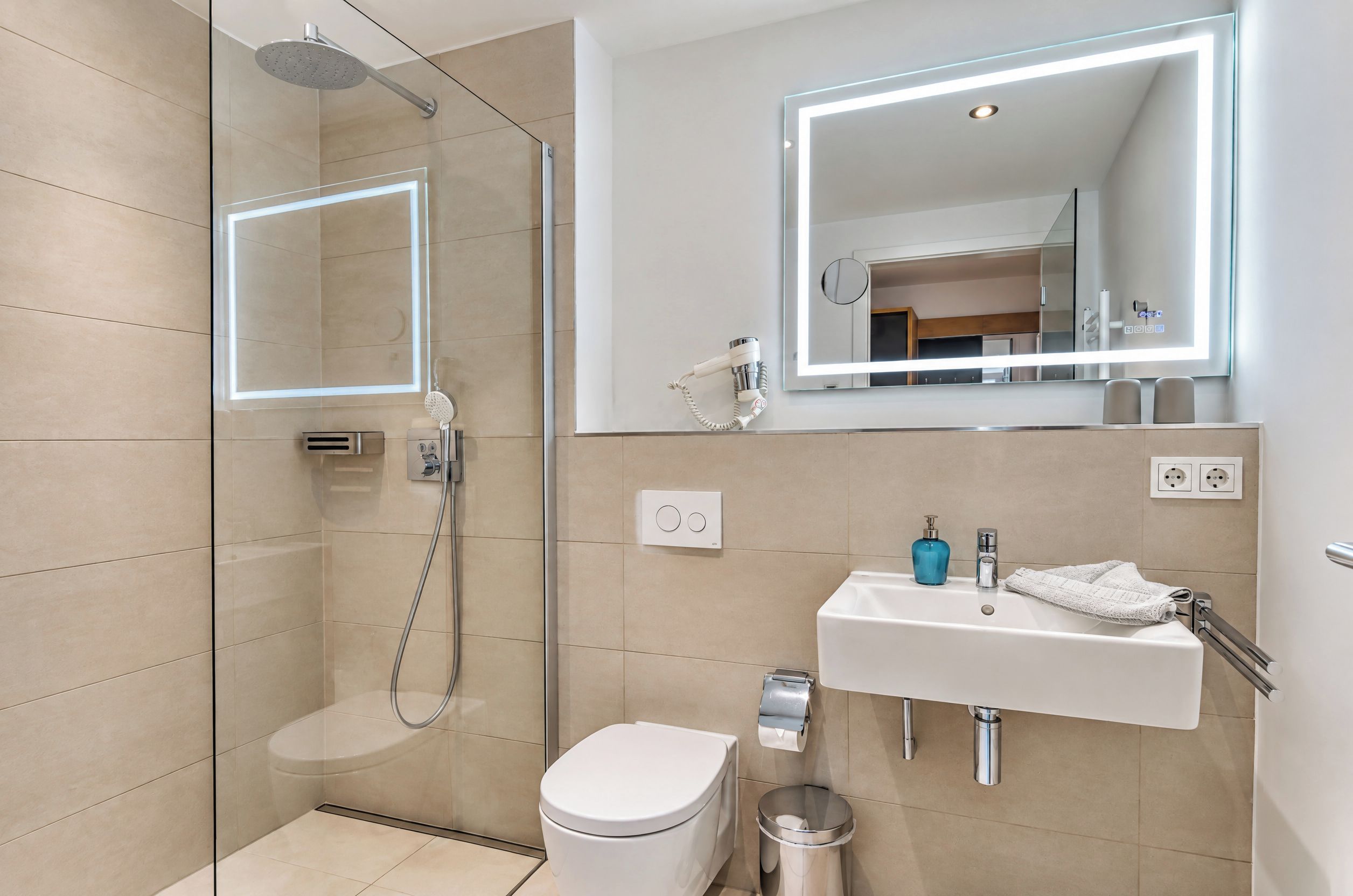 Ein modernes Badezimmer mit einer in Glas abgeschlossenen Dusche, einer weißen Toilette, einem Waschbecken mit blauem Seifenspender und einem Spiegel mit integrierter Beleuchtung.