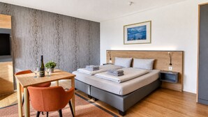 A modern bedroom with a bed, a table with a bottle and glasses, a chair, a small plant, and a framed picture on the wall.