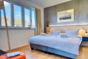A bedroom with a large bed, a window with curtains, a framed picture on the wall, and a bedside table with a lamp.