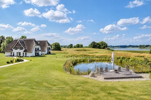 Een huis met rieten dak en een vijver met terras en stoelen.
