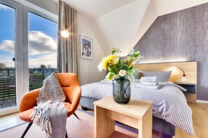 Une chambre moderne avec une grande fenêtre, un lit recouvert d’une couette à carreaux, une table de chevet ornée d’un vase de fleurs et un fauteuil orange.