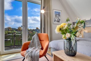 Une chambre moderne avec une grande fenêtre, un fauteuil orange confortable, une table de chevet en bois et un vase rempli de fleurs.