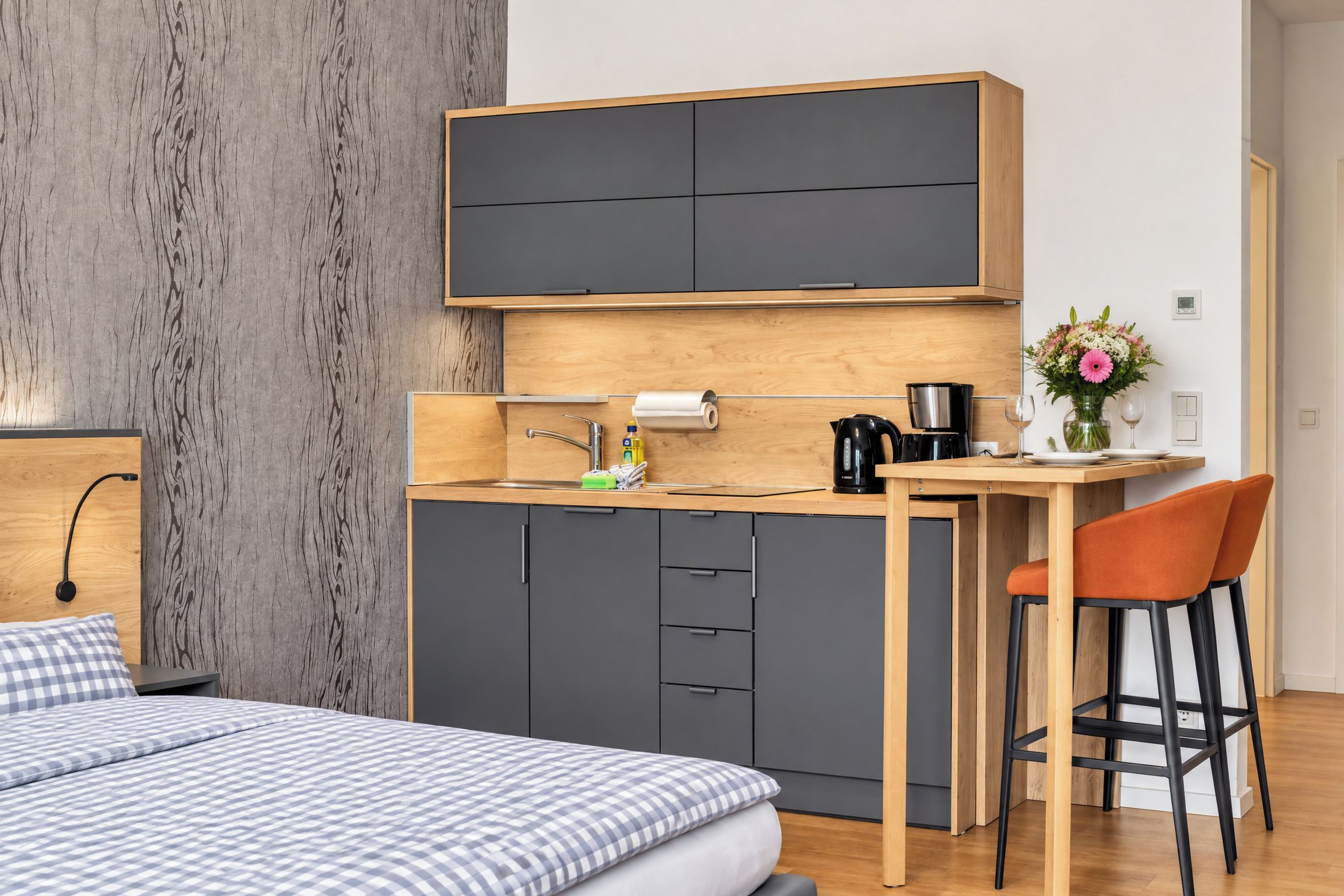 Un dormitorio compacto con cama, una kitchenette pequeña con cafetera y una zona de comedor con una silla.