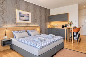 A modern bedroom with a large bed, a small desk, and a kitchenette.