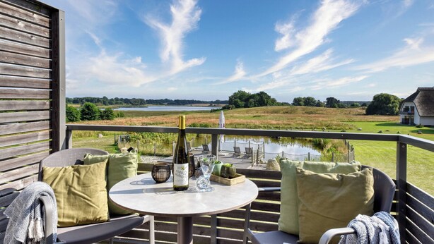 A balcony with a table, chairs, and a bottle of wine. The balcony overlooks a scenic landscape with a body of water and trees. - Vacation apartment Wiesenpieper in Wreecher Idyll (Putbus)