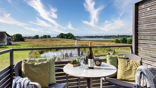 A balcony with a table, chairs, and a bottle of wine, overlooking a lake and fields.