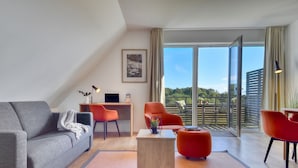 Un salon moderne avec un canapé gris, des fauteuils rouges, une table basse en bois et une grande porte-fenêtre coulissante donnant sur un balcon avec vue sur la verdure.