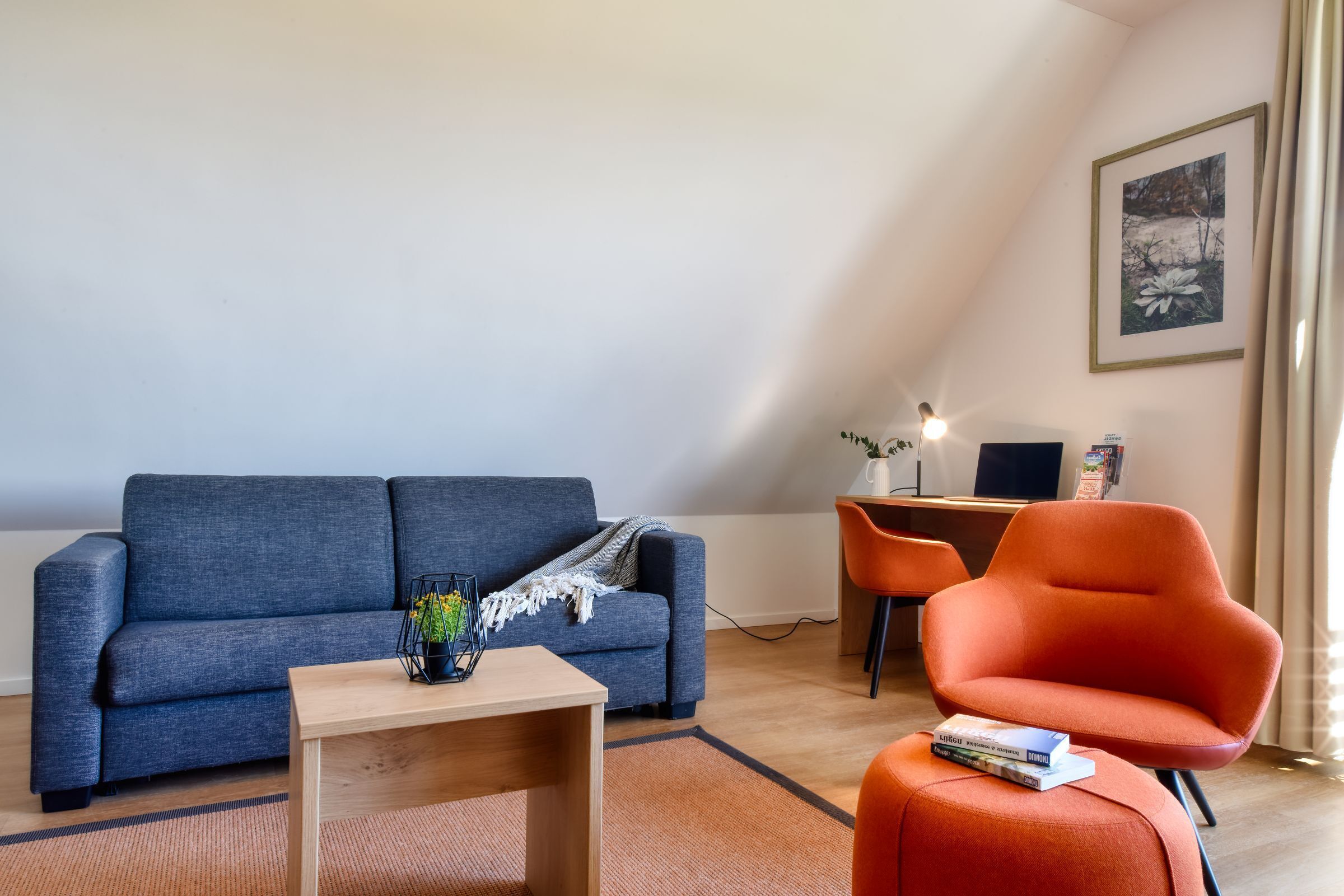 Un salon moderne avec un canapé gris, une table basse en bois et un fauteuil orange. On y trouve un bureau avec un ordinateur et une lampe.
