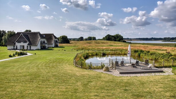 A large house with a pond and a deck with chairs and a table.