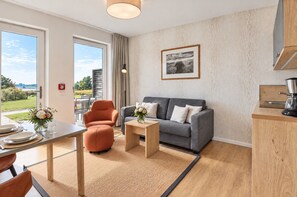 A modern living room with a grey sofa, orange armchair, and a small coffee table. There's a dining area with a table set for two, and a kitchen area with a coffee machine. The room has large windows offering a view of the outdoors. - Vacation apartment in the idyllic village of Wreecher on the shores of Lake Wreechen (Putbus)