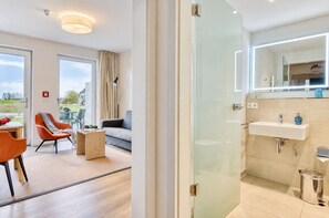A modern hotel room with a glass door leading to a balcony, a seating area with a sofa and chairs, and a bathroom with a sink and mirror.