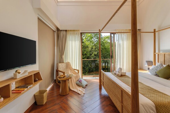 Un dormitorio con una cama grande, un televisor, una silla de madera, una mesita y una ventana con vistas a la vegetación.