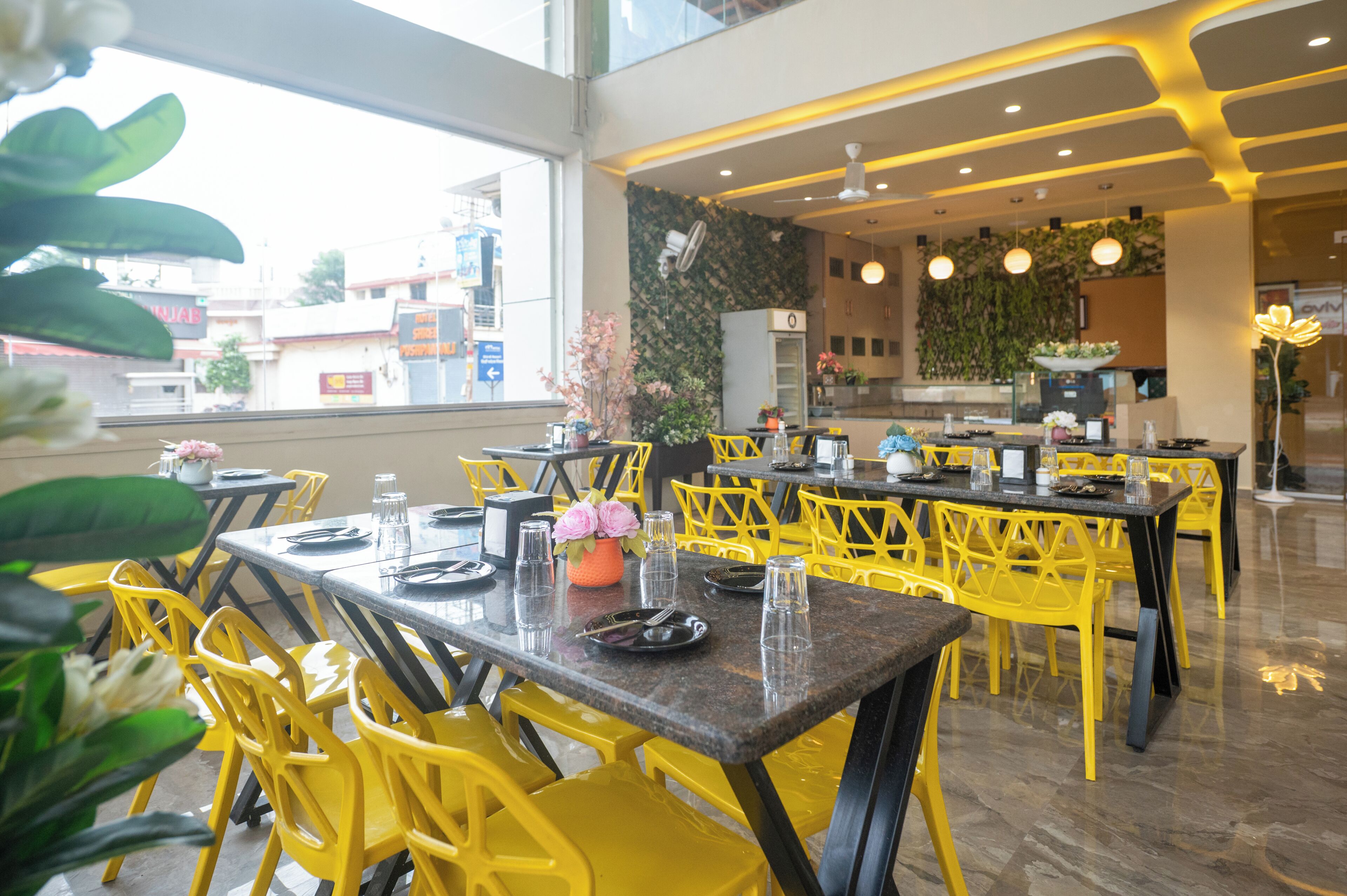 A restaurant with yellow chairs and tables, a kitchen area, and a view of buildings outside.