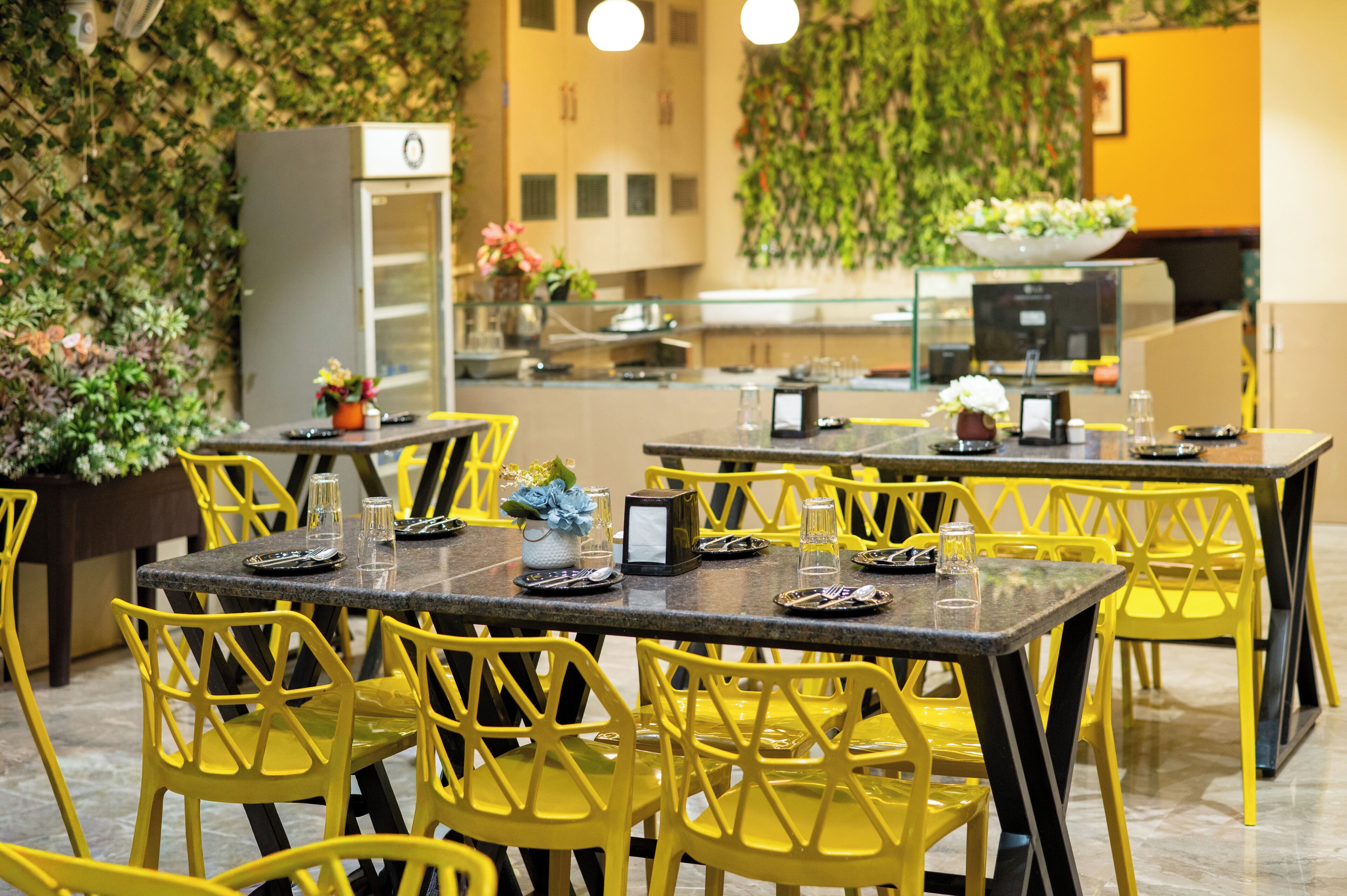 A restaurant with yellow chairs and tables, a kitchen area, and a green wall with plants.