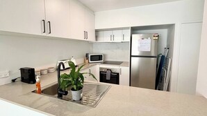 Eine moderne Küche mit weißen Schränken, einem Edelstahlkühlschrank, einer Mikrowelle, einem Wasserkocher und einem Spülbecken mit einer Pflanze.