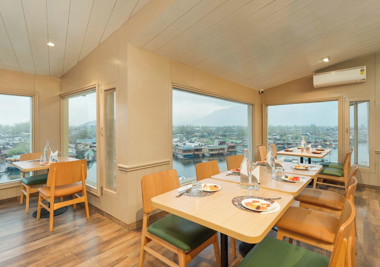 A dining area with wooden tables and chairs, large windows with a view of boats and a mountain, and an air conditioning unit on the wall.