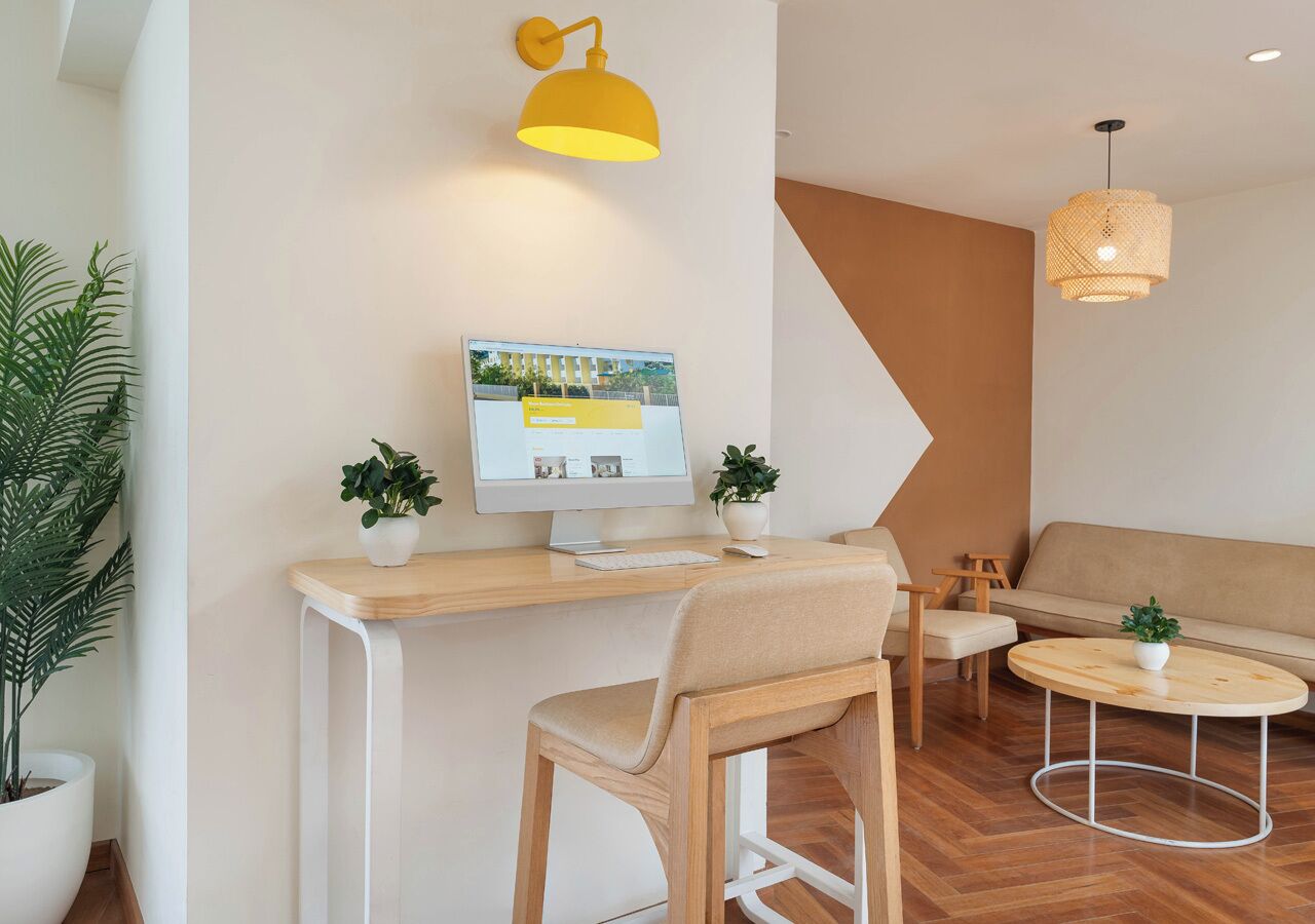 A modern living room with a wooden desk, a computer, two potted plants, a round coffee table, and a beige sofa.