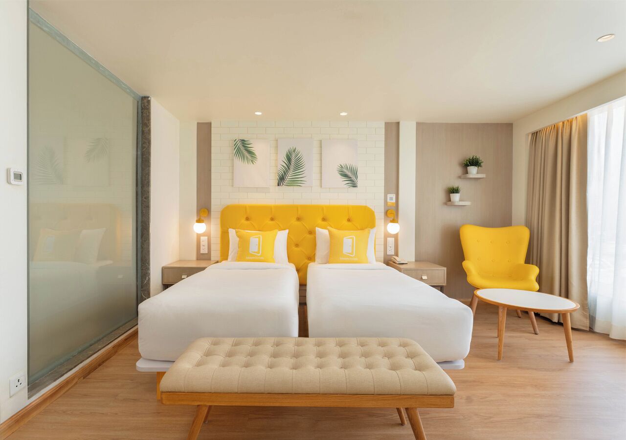 A hotel room with a double bed, a small table, a yellow armchair, and a large window with curtains.