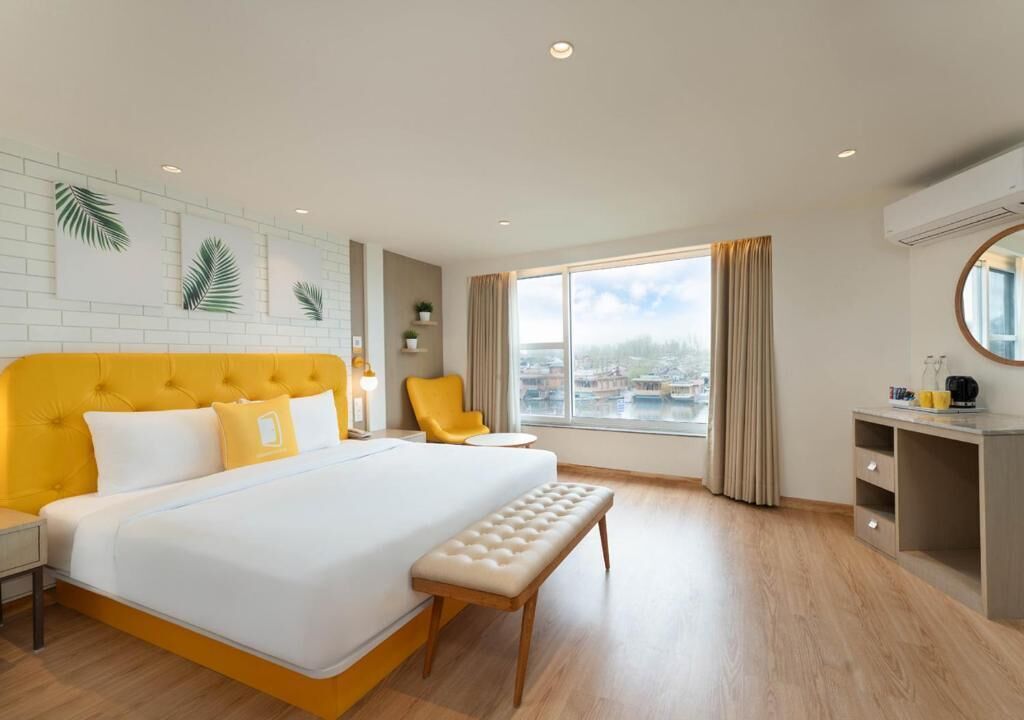 A hotel room with a large bed, a bench, a desk, and a view of a cityscape through the window.