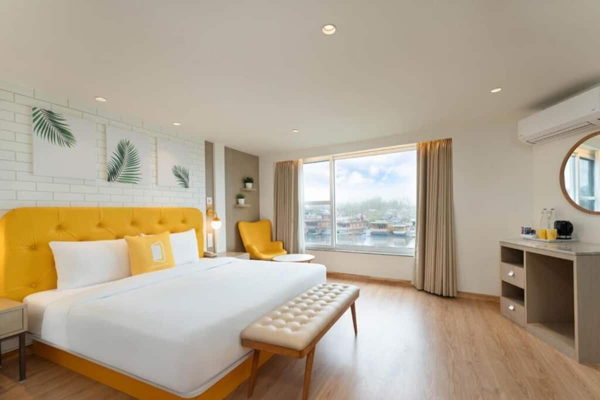 A hotel room with a large bed, a bench, a desk, and a view of a cityscape through the window.