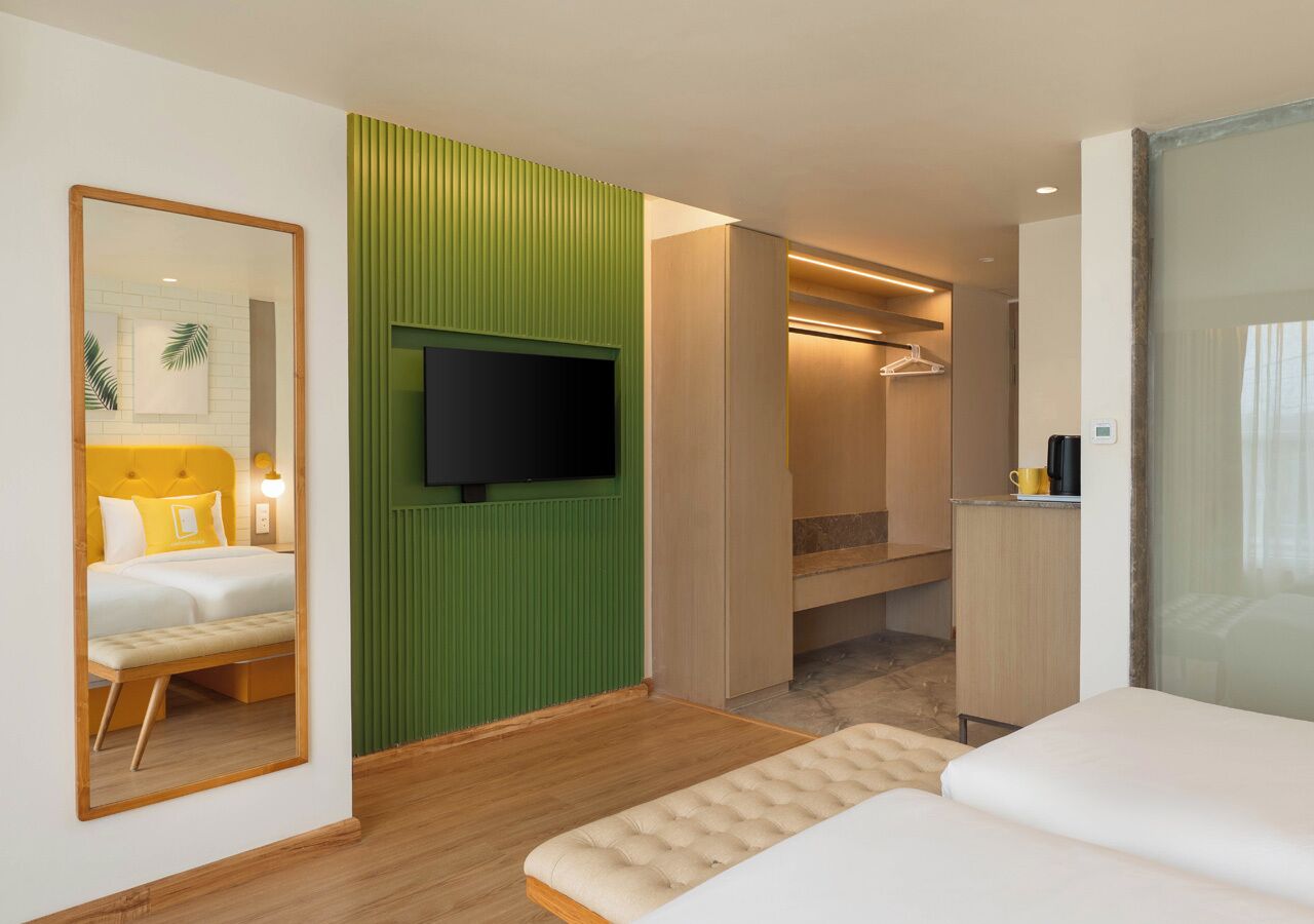 Quarto de hotel com cama, televisão fixada numa parede verde, chão de madeira e banheiro visível através de uma porta aberta.
