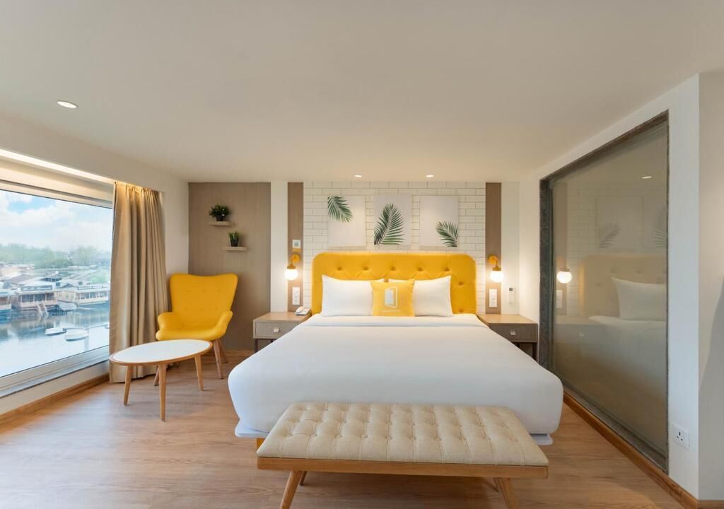 A hotel room with a large bed, a yellow armchair, a small table, and a view of a cityscape through a large window.