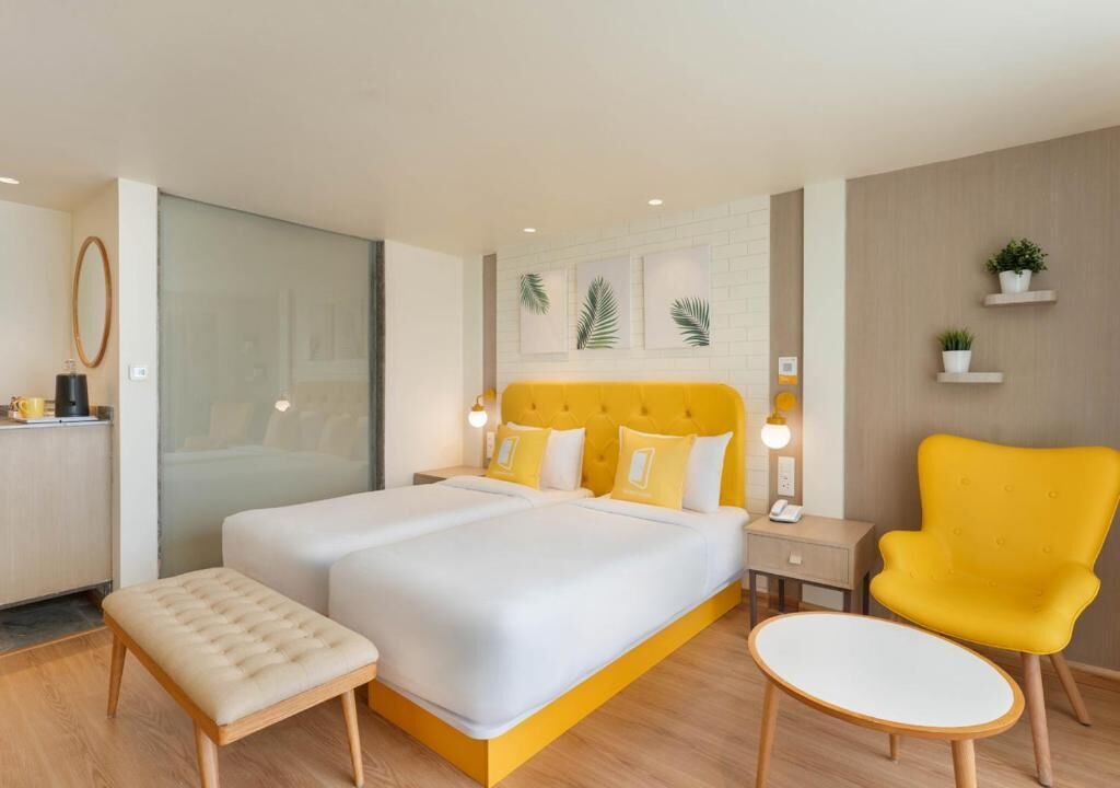 A modern hotel room with a large bed, a small stool, a yellow armchair, and a bedside table with a lamp.