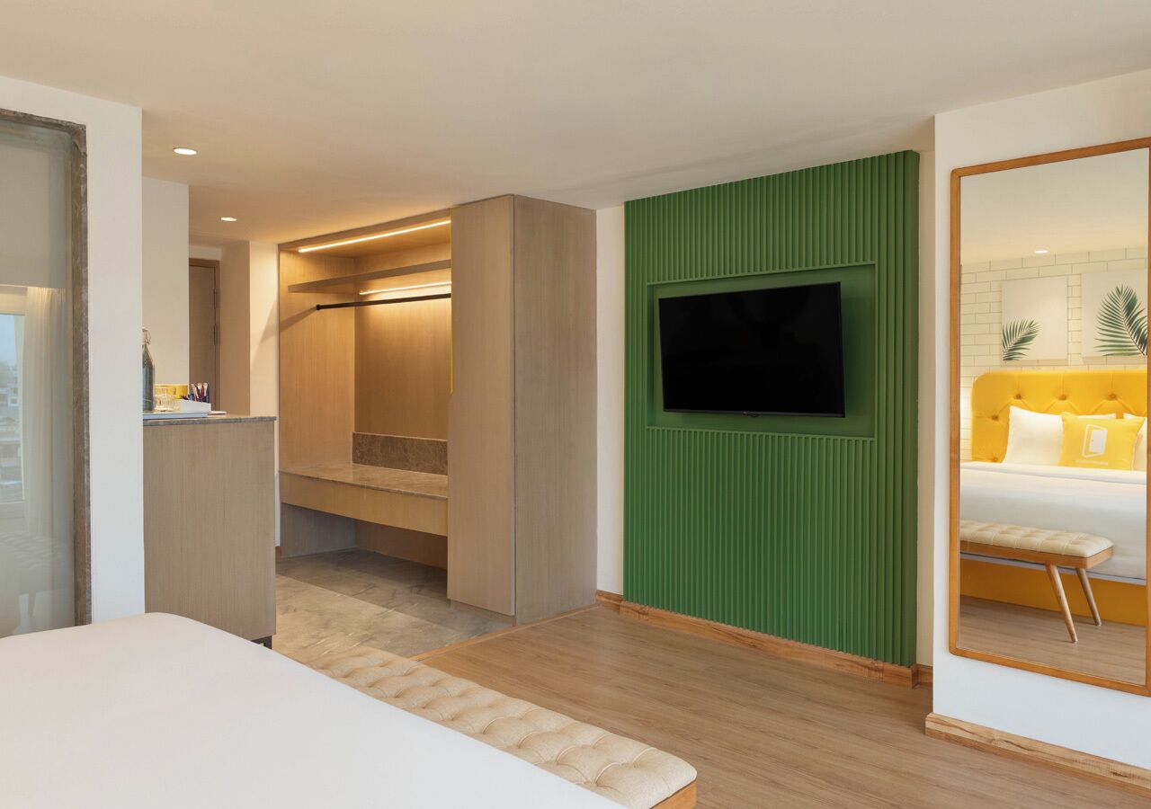 A modern hotel room with a bed, a TV mounted on a green wall, a wooden wardrobe, and a small kitchenette.