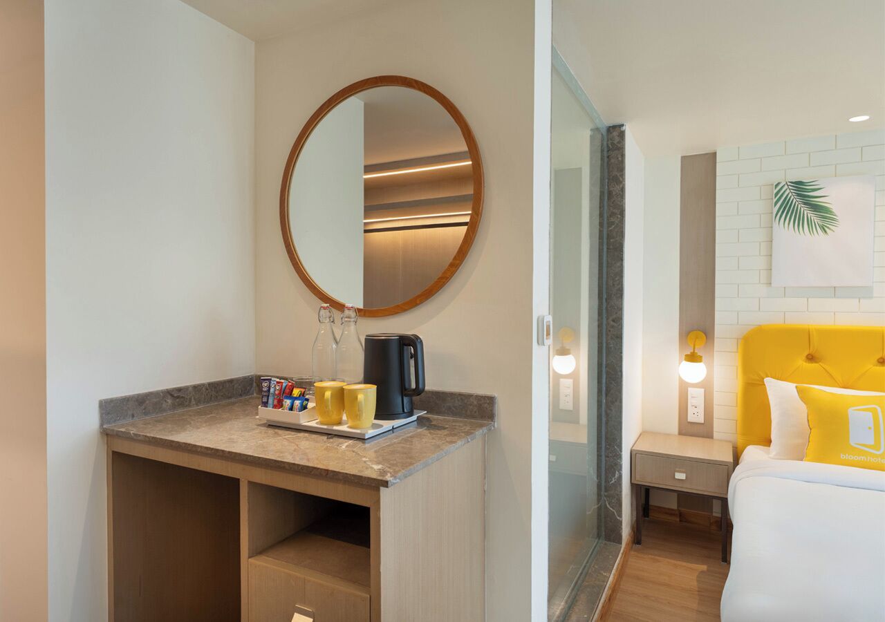 Quarto de hotel com secretária de madeira, espelho redondo, cama com cabeceira amarela e luminária fixada na parede.