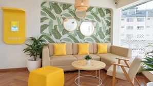 A modern hotel lobby with a sofa, round coffee table, and yellow ottoman. A large mirror and a check-in sign are visible. - Bloom Boutique - Dal Lake (Srinagar)