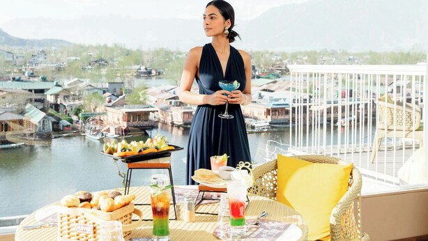 A balcony with a table set for a meal, a person holding a drink, and a view of a river and buildings in the background. - Bloom Boutique - Dal Lake (Srinagar)