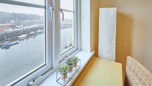 A room with a window overlooking a harbor, a wooden table, and a white textured wall.