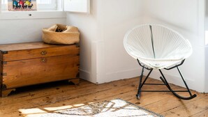 A wooden chest of drawers, a woven basket, a white rocking chair, and a patterned rug in a room with a wooden floor and a window. - Central 3 Bedroom Apartment in Ericeira (Mafra)