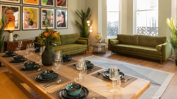 Un coin repas avec une longue table dressée pour six personnes, des canapés verts et des œuvres d’art encadrées accrochées aux murs.