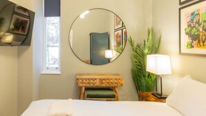 Un lit bien fait, avec une serviette pliée, un miroir rond, une armoire en bois avec un coussin vert, une plante en pot et un tableau encadré accroché au mur.