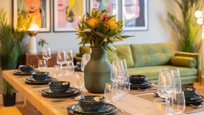 Une table à manger dressée avec des assiettes, des bols et des verres à vin, entourée de canapés verts et ornée d’œuvres d’art encadrées au mur.