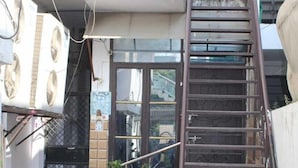 A residential building with a metal staircase leading to a balcony. There are potted plants and a small garden area in front of the building.