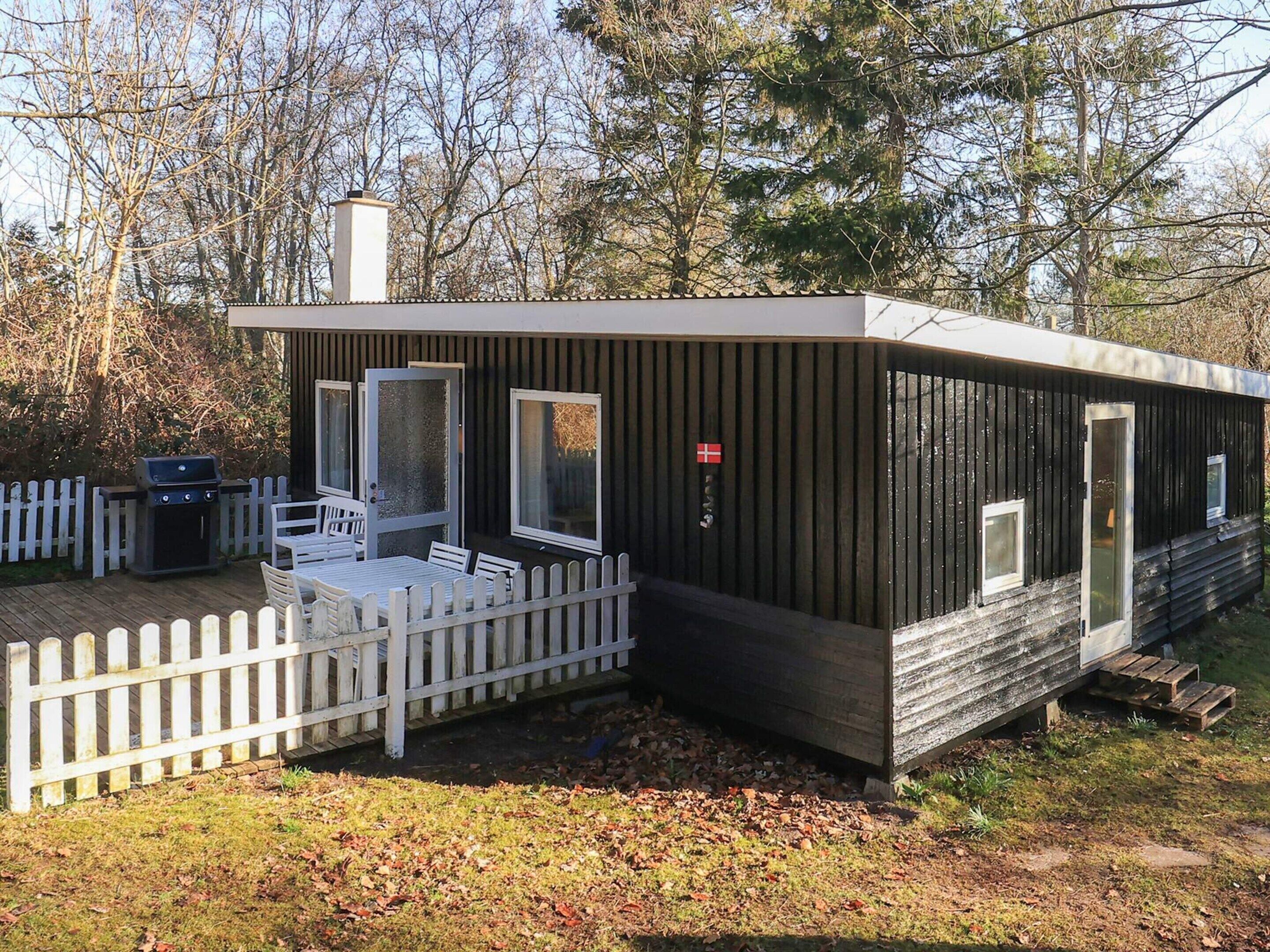 Une petite cabane à un seul étage, avec une clôture blanche en bois, un barbecue et une porte ornée d’un drapeau.